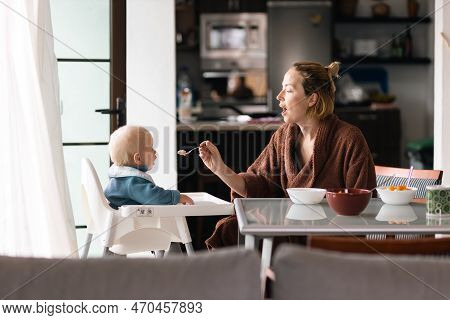 Mother Wearing Bathrope Spoon Feeding Her Infant Baby Boy Child Sitting In High Chair At The Dining 