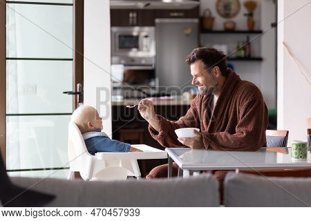 Father Wearing Bathrope Spoon Feeding Hir Infant Baby Boy Child Sitting In High Chair At The Dining 