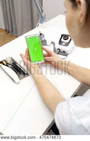 Manicure, Pedicure Technician Holds Smartphone With Green Chroma Key Screen, Sitting At Workstation 