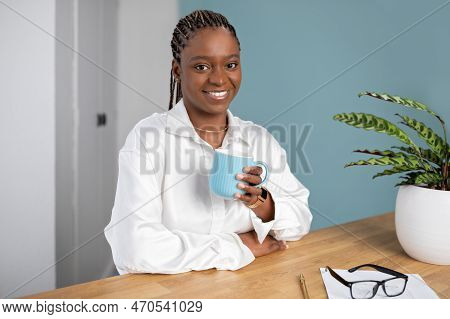 Positive Young Black Woman Sitting At Workdesk, Drinking Coffee