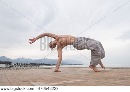 Shirtless Caucasian Man Doing Backflip On Pebble Beach.