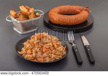 Stewed Cabbage - Bigos In A Black Plate Against The Background Of A Circle Of Smoked Sausage And Bak