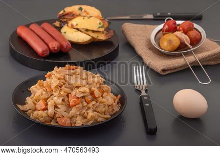Stewed Cabbage - Bigos In A Black Plate Against The Background Of Pickled Vegetables And Baked Potat