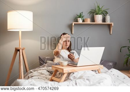 Indoor Shor Of Sick Ill Woman Wearing Sleeping Mask Holding Laptop While Lying On Bed, Using Cell Ph