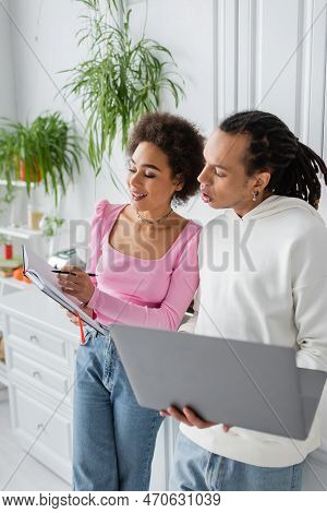 Cheerful African Amercan Woman Holding Notebook Near Boyfriend And Laptop In Kitchen.
