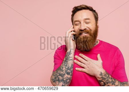 Happy Tattooed Man With Closed Eyes Touching Chest While Talking On Smartphone Isolated On Pink.