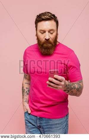 Tattooed Man In Magenta T-shirt Standing With Hand In Pocket Of Jeans And Using Smartphone Isolated 