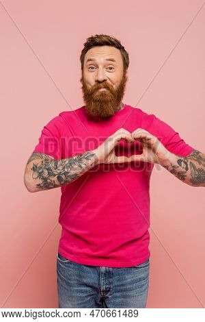 Cheerful Tattooed And Bearded Man In Magenta T-shirt Showing Heart Sign While Looking At Camera Isol