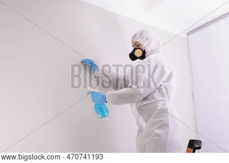 Woman In Protective Suit Cleaning Mold With Sprayer On Wall Indoors