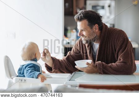 Father Wearing Bathrope Spoon Feeding Hir Infant Baby Boy Child Sitting In High Chair At The Dining 