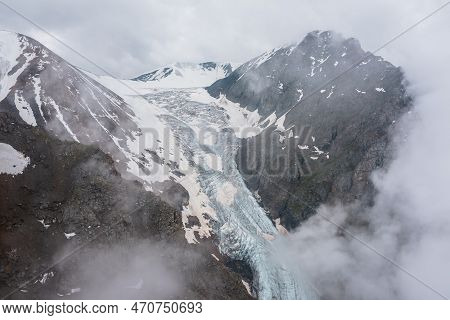 Atmospheric Mountain Landscape With Long Vertical Glacier Tongue With Cracks Among Rocks In Dense Lo