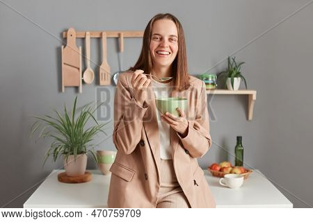 Indoor Shot Of Smiling Laughing Young Adult Woman Wearing Beige Suit Standing Near Table On Kitchen 