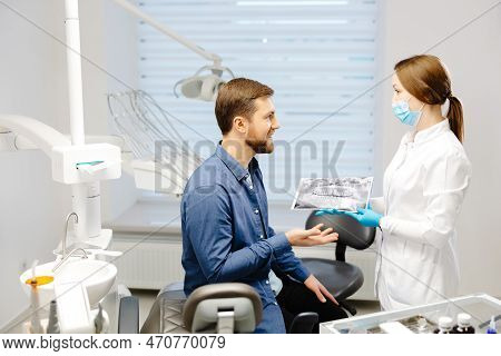 Young Attractive Man Visiting Dentist, Sitting In Dental Chair At Modern Light Clinic. Young Woman D