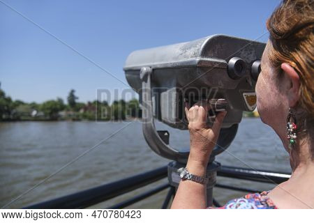 Latin Mature Woman Viewing Through Public Binoculars On The Lujan River Bank In Tigre, Buenos Aires,