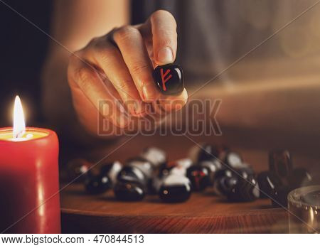 Man's Hand Holds A Rune Of Fehu With Fingers. He Shows A Rune. In Front Of Him On A Wooden Tray Ston