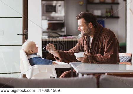 Father Wearing Bathrope Spoon Feeding Hir Infant Baby Boy Child Sitting In High Chair At The Dining 