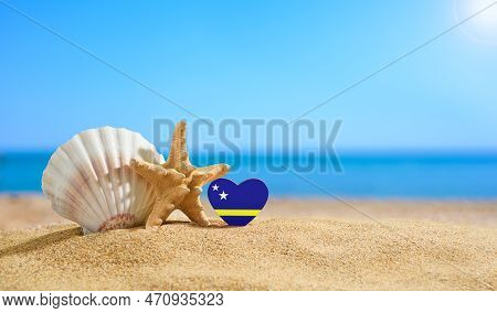 Beautiful Beach In The Curacao. Flag Of Curacao In The Shape Of A Heart And Shells On A Sandy Beach.