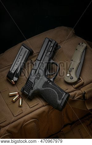 A Pistol, Cartridges For It And A Folding Knife On A Brown Backpack. Self-defense And Survival Kit. 