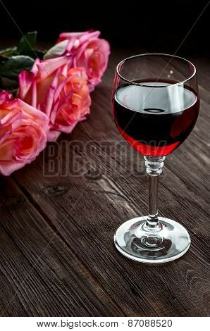 A glass of red wine and three roses on wooden background.