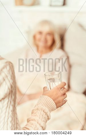 Time to return love doubly. Happy thankful young woman standing in the bedroom and taking care of aged mother while holding glass of water