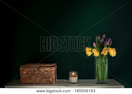 yellow daffodil flowers with pupple tulip blooming in vase with green wall. Dark key. Interior with candle