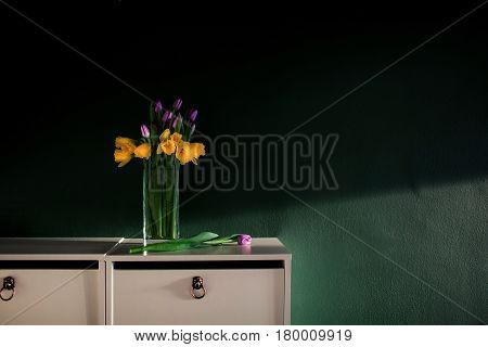 yellow daffodil flowers with pupple tulip blooming in vase with green wall. Dark key. Interior with candle