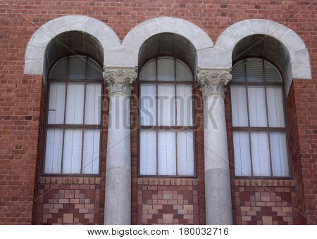 Red brick wall with three recessed arched paned windows beneath a trim of three white arches and separated by two white columns