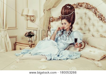 Indoors shot in the Marie Antoinette style. A young sexy girl in a lush blue retro dress with a high hairstyle lies on the bed with a glass of sparkling wine in her hand. Woman tired of luxury.