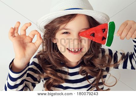 Little girl with big smile is showing OK by her fingers and covering her eye with a lollipop watermelon shaped. Image is on a white studio background. Good mood concept
