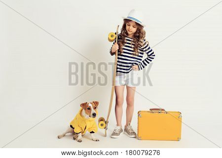 Young kid holding longboard is standing with dog which is dressed in jacket. Image on a white studio background. Vacation, holidays, summer time, voyage concept