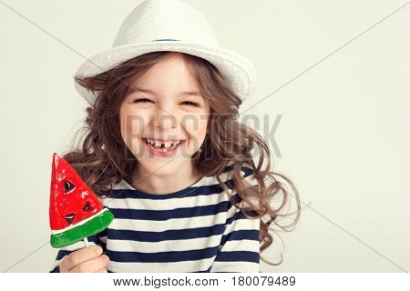 Girl with curly hair is smiling from ear to ear and holding a lollipop watermelon shaped. Picture is taken a studio and has white background. Childhood concept