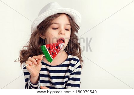 Young girl wearing white hat is licking a huge lollipop watermelon shaped with a pleasure and concentration at the same time.
