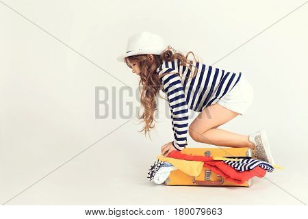 Little girl is trying to lock up overflowing suitcase by sitting on it. A picture is taken at studio and has white background