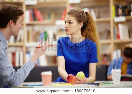 Portrait of pretty teenage girl talking to groupmate at table in library hall, discussing student project and eating lunch