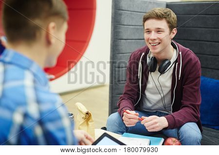 Portrait of smiling teenage boy talking to friend while sitting at table with books and lunch in college