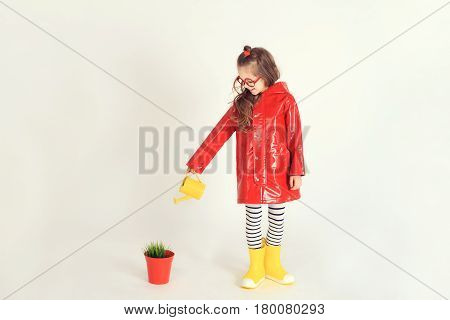 Young girl wearing raincoat and rubber boots is watering grass from a pot by using watering can. The picture is taken at studio and has white background