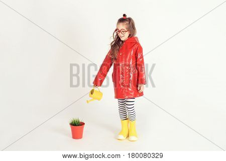 Smiling girl wearing raincoat and rubber boots is watering grass from a pot by using watering can. The picture is taken at studio and has white background