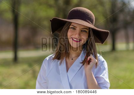 Portrait of a smiling young woman wearing a hat in a park during spring. Woman bowed her head head to the right side of frame. Medium shot. Shallow depth of field.