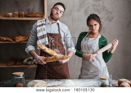 Young man with two loafs and woman with rollin pin standing in kitchen earnestly