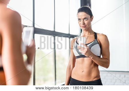 Portrait of pretty brunette woman smiling taking mirror selfie in gym,  posing boasting her fit figure and slim waist