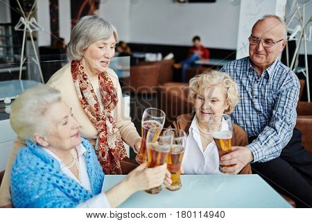 Having good time with friends in pub: group of joyful senior people sitting at table and clanging glasses of beer together