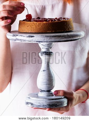 Woman's hands hold a cheesecake on a wooden cake stand.Cheesecake decorated with cherry sauce with berries