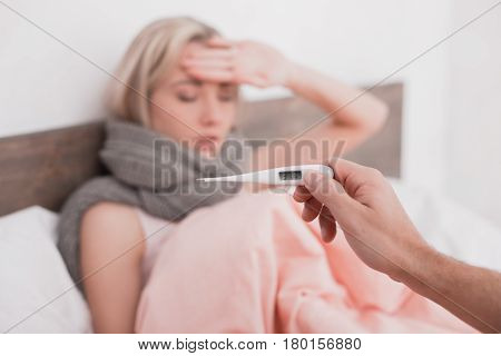 Young woman sitting on bed with fever while man holding thermometer with a high tempreture