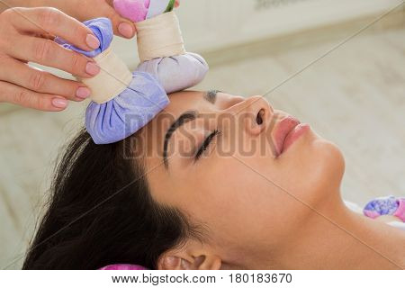 Herbal ball face massage in ayurveda spa. Female massagist with young woman in wellness center. Healthcare therapy to beautiful indian girl in beauty parlor, side view of face with eyes closed