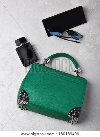 Nice beautiful small womans leather green fashionable bag with decorative metallic spikes or thorns on white textured background near sunglasses perfume and case