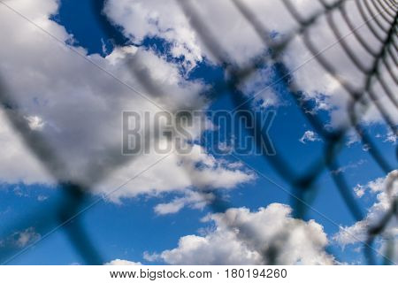 Background of Metal mesh fence against deep blue sky with white fluffy clouds backdrop. Concept of jail and freedom.