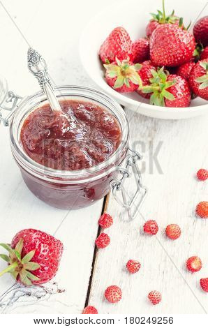 Homemade delicious strawberry jam in a glass jar and fresh strawberries on white rusric wooden background. Selective focus.