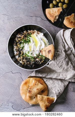 labneh middle eastern lebanese cream cheese dip with olive oil, salt, herbs, olives tapenade served in black bowl with traditional pita bread over gray texture metal background. Top view with space