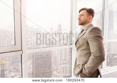 Serious work. Professioanl confident businessman standing near window and holding his hands in the pockets while being involved in thoughts