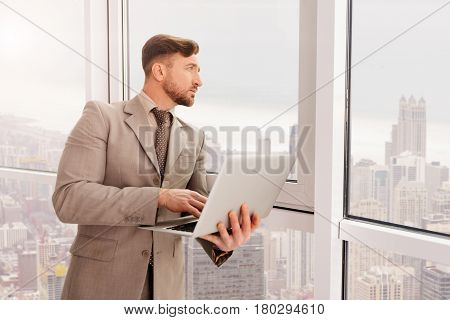 What is outside. Professioanl serious businessman using laptop and standing near window while enjoying the view on the city